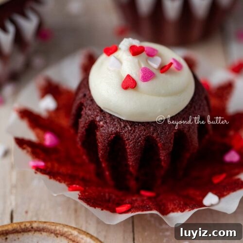 Opened red velvet cupcake with cream cheese frosting topped with heart sprinkles on a light wood background.