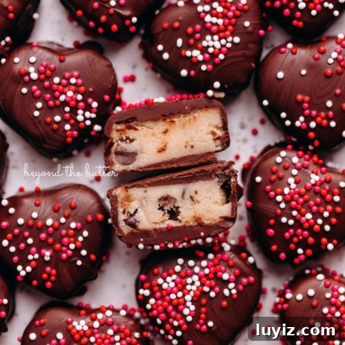 Chocolate covered cookie dough hearts topped with red, pink, and white sprinkles placed on a white parchment paper lined baking sheet with center heart cut in half to see the cookie dough inside.