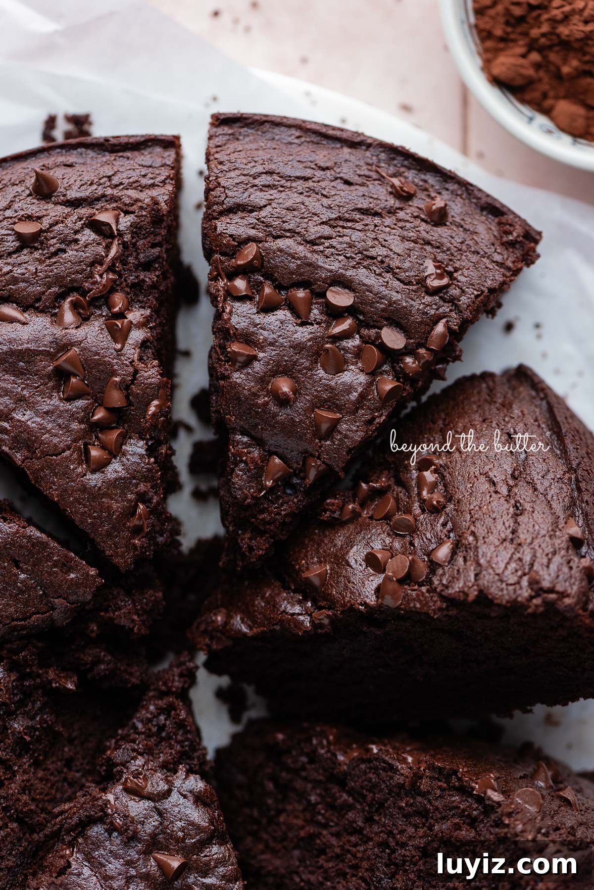 Decadent Chocolate Ricotta Cake Recipe for Moist, Fluffy Results 3 Slices of chocolate ricotta cake topped with chocolate chips on white parchment paper and a small bowl of cocoa powder placed on a light pink tiled background.
