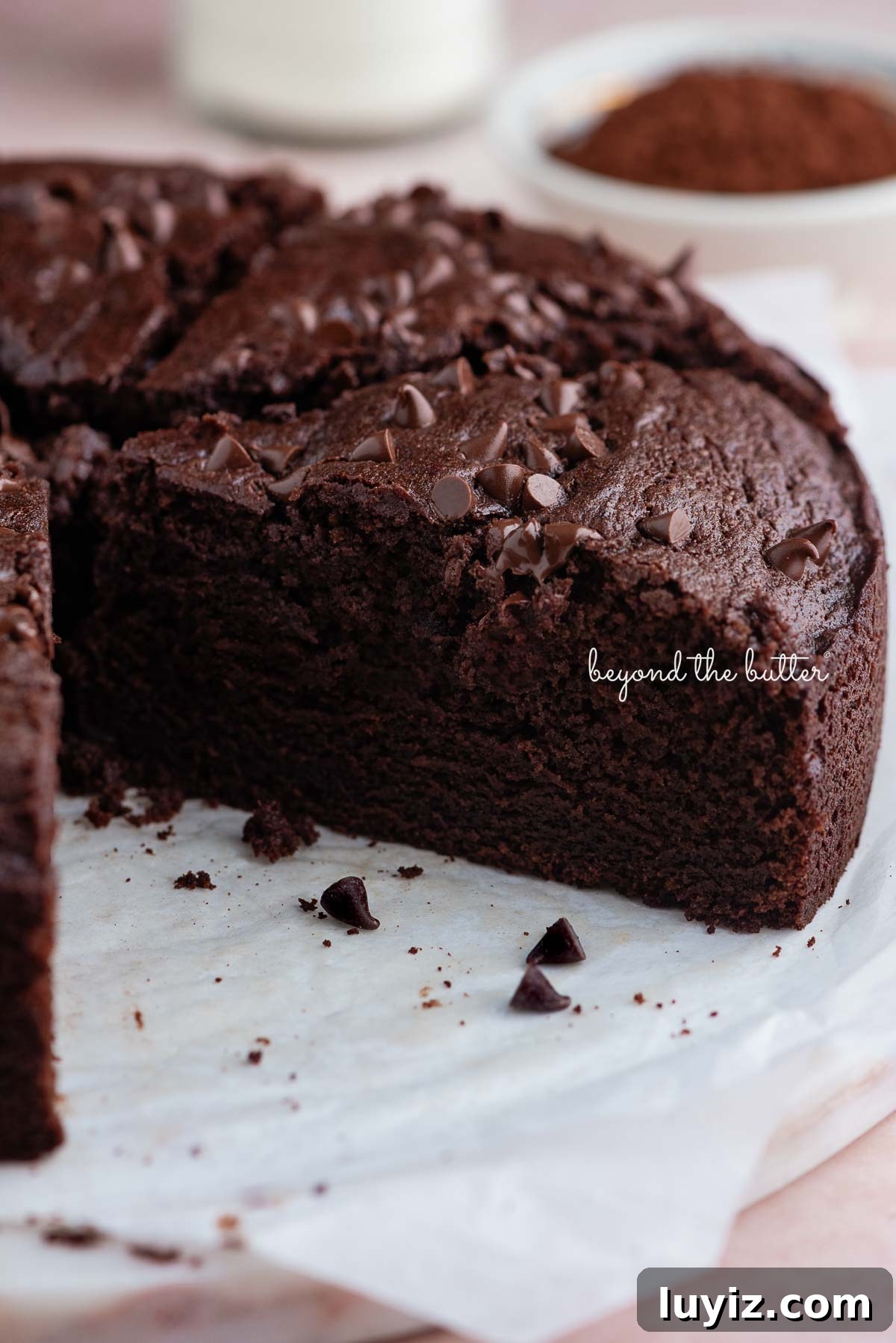 Decadent Chocolate Ricotta Cake Recipe for Moist, Fluffy Results 10 Slices of chocolate ricotta cake on white parchment paper and a white marble cake plate placed on a pink tiled background with a glass of milk and small bowl of cocoa powder nearby.