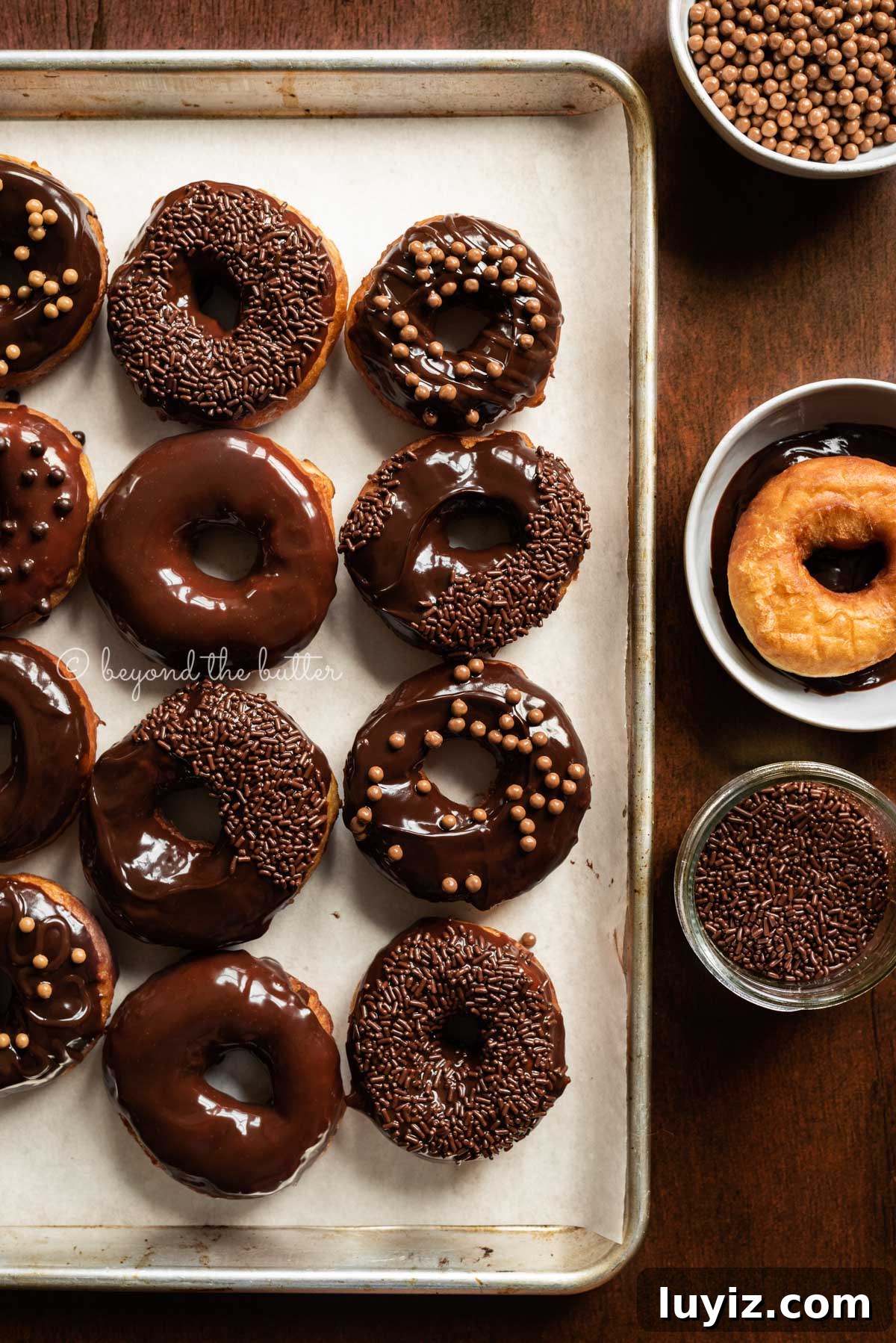 Homemade Donuts from Scratch: Classic Recipe for Soft, Fluffy Donuts 7 Chocolate glazed decorated donuts on a parchment paper lined baking sheet with a donut and chocolate jimmies next to them | All Images © Beyond the Butter™
