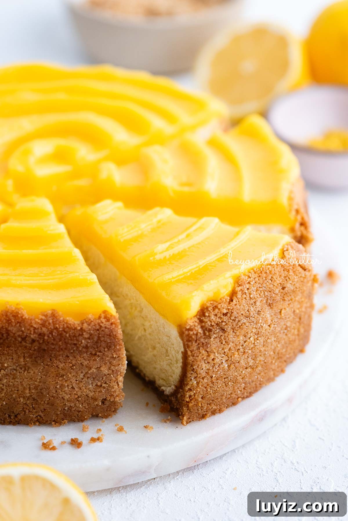 Creamy Lemon Cheesecake Recipe with Zesty Citrus Glaze 13 Slice of lemon cheesecake topped with lemon curd being removed from the cake plate with cake server on a white background.