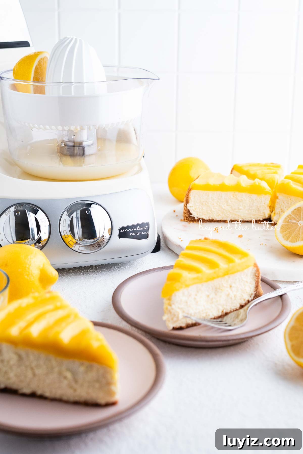 Creamy Lemon Cheesecake Recipe with Zesty Citrus Glaze 3 Slices of lemon cheesecake topped with lemon curd on pink dessert plates with cream colored mixer on white background.