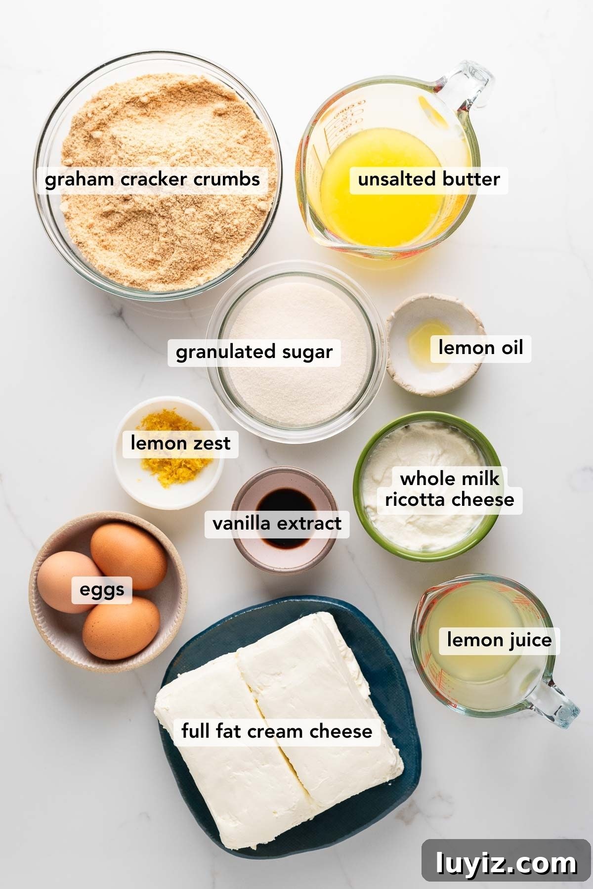 Creamy Lemon Cheesecake Recipe with Zesty Citrus Glaze 4 Labeled ingredients in bowls used to make lemon cheesecake on a white marbled background.
