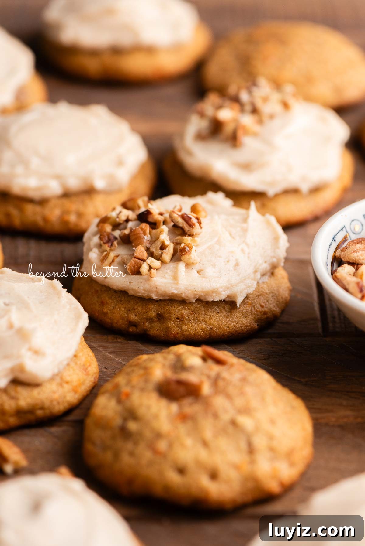 Carrot Cake Cookie Recipe: Moist, Spiced Cookies with Cream Cheese Frosting 11 Carrot cake cookies frosted with brown butter cream cheese frosting and some topped with chopped pecans with a small bowl filled with chopped pecans to the side on a dark wood background.