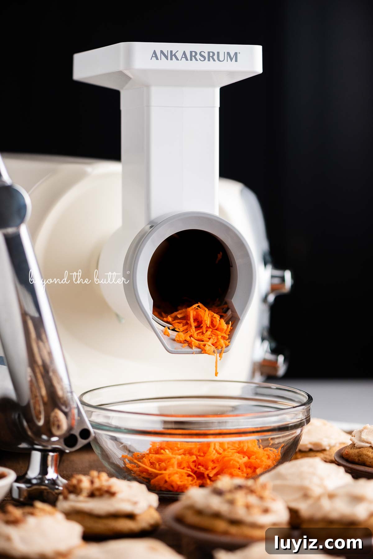 Carrot Cake Cookie Recipe: Moist, Spiced Cookies with Cream Cheese Frosting 3 Light creme color ankarsrum mixer using the vegetable cutter attachment to grate carrots into a glass bowl for carrot cake cookies against a dark background.
