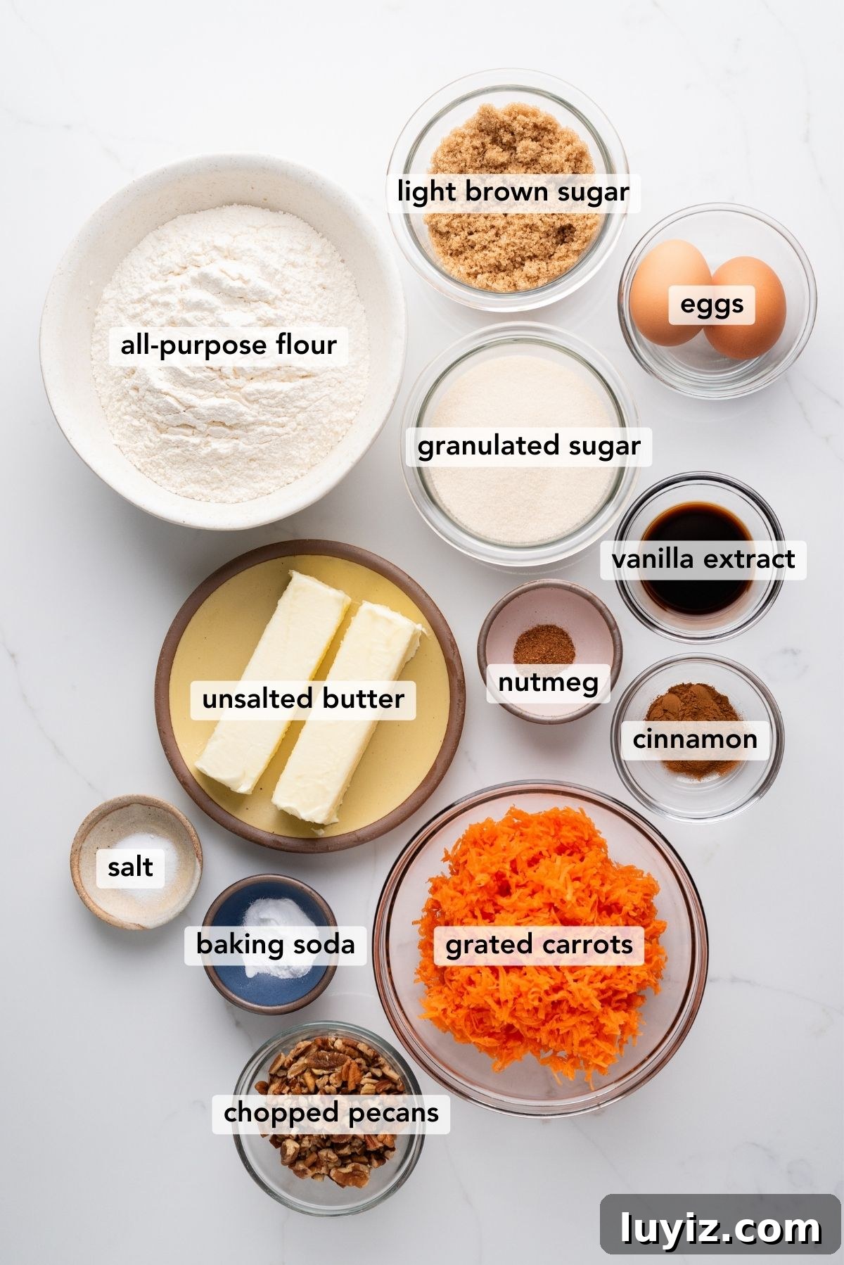 Carrot Cake Cookie Recipe: Moist, Spiced Cookies with Cream Cheese Frosting 4 Carrot cake cookies ingredients in various size bowls labeled as all-purpose flour, baking soda, cinnamon, nutmeg, salt, unsalted butter, light brown sugar, granulated sugar, eggs, vanilla extract, grated carrots, and chopped pecans on a white marbled background.