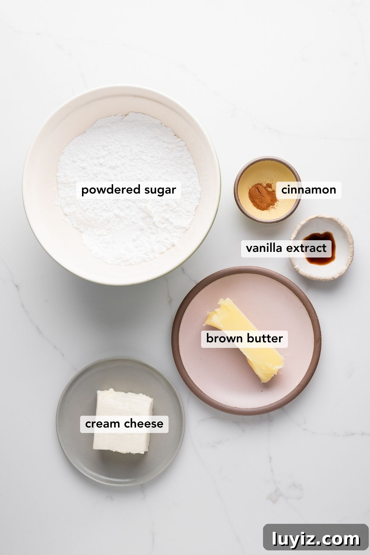 Carrot Cake Cookie Recipe: Moist, Spiced Cookies with Cream Cheese Frosting 5 Brown butter cream cheese frosting ingredients in various size bowls labeled as powdered sugar, cinnamon, vanilla extract, brown butter, and cream cheese on a white marbled background.