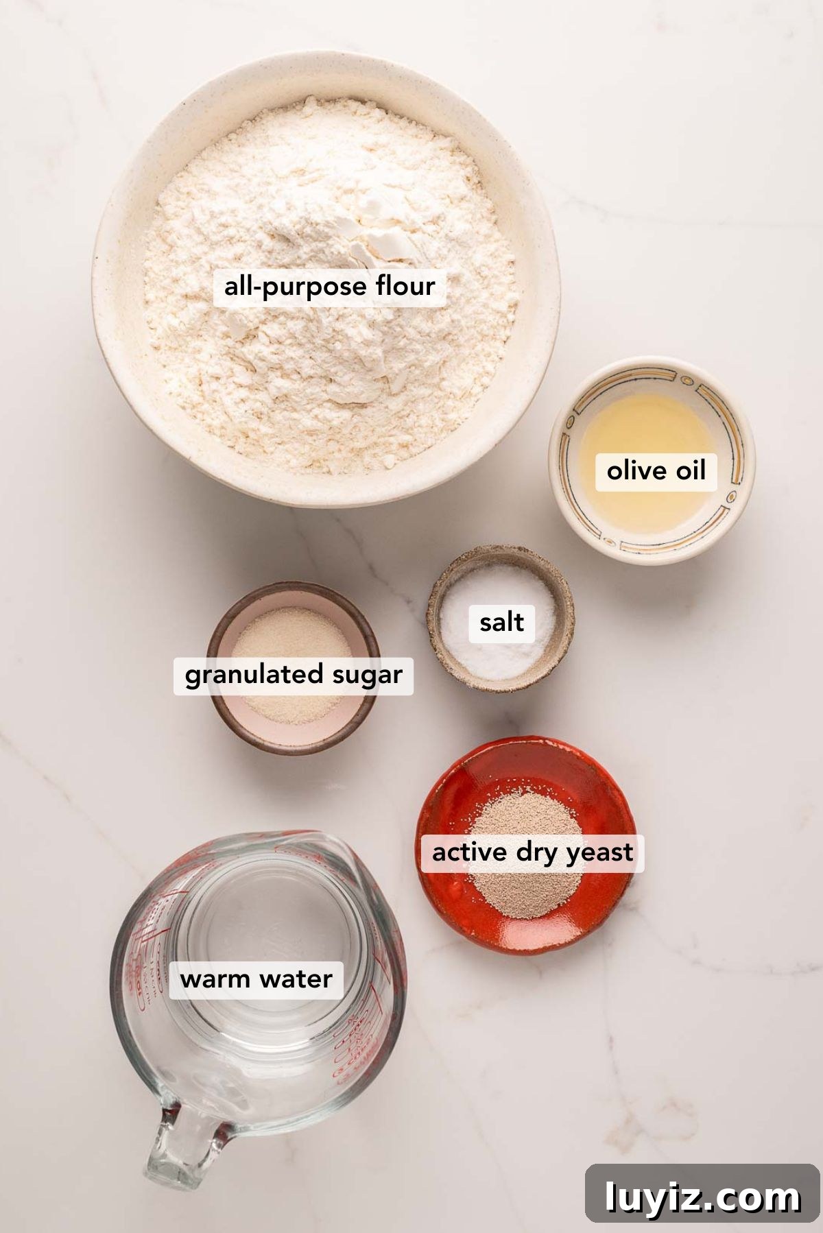 Classic French Bread Recipe for Crispy Crust and Soft Crumb 3 Ingredients in various size bowls labeled as all-purpose flour, olive oil, granulated sugar, salt, warm water, and active dry yeast on a white marbled background.