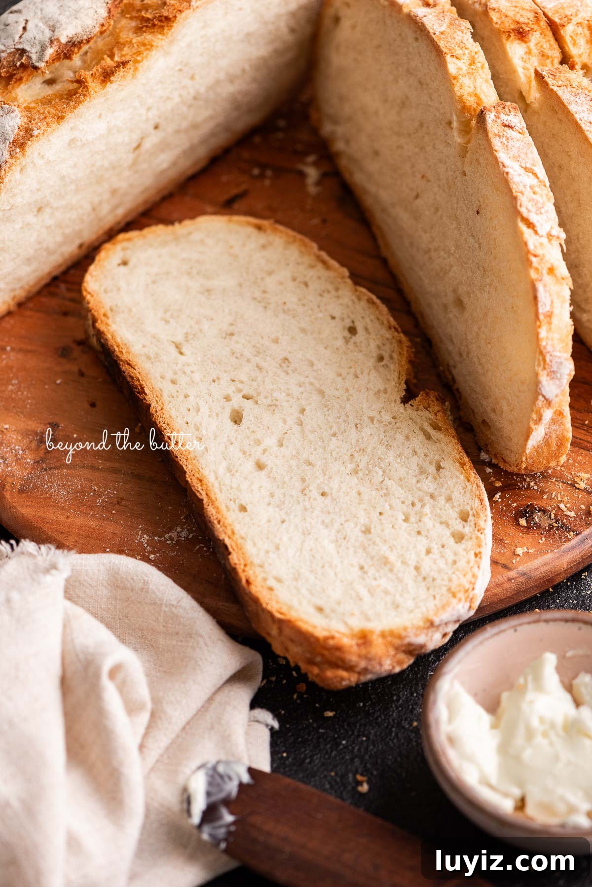 Classic French Bread Recipe for Crispy Crust and Soft Crumb 10 Sliced french bread placed on a round wooden cutting board next to a small pink bowl of butter and a light beige cloth napkin on a black background.