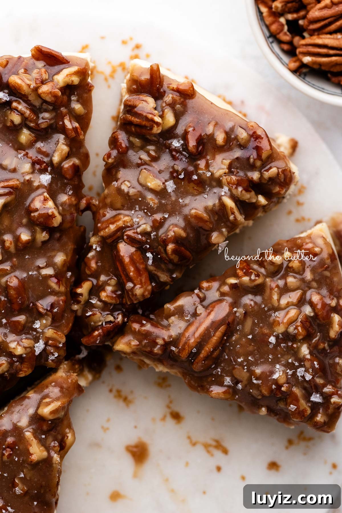 Pecan Pie Cheesecake Recipe — Decadent Homemade Dessert 15 Slices of pecan pie cheesecake topped with sea salt flakes on a white marble cake stand next to a small bowl of pecans on a white background.