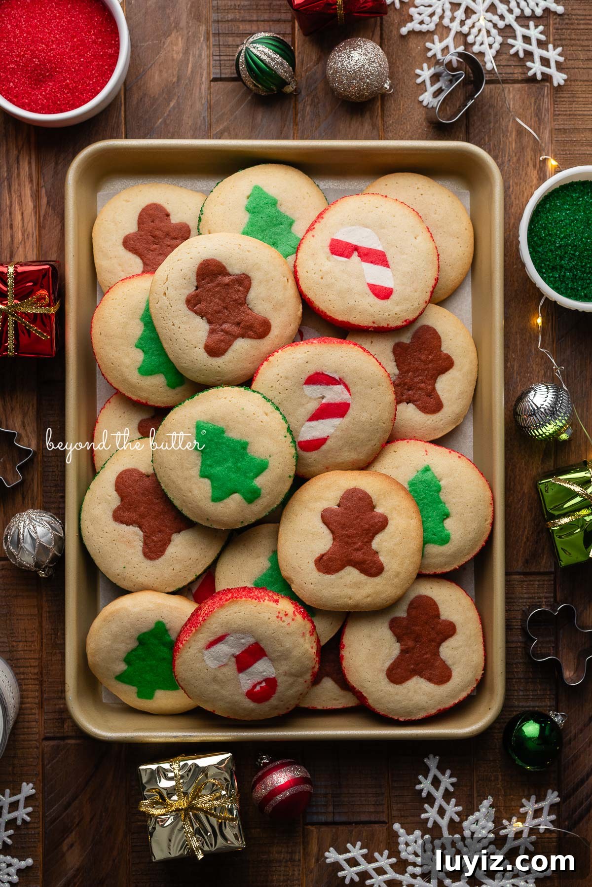 Festive Slice-and-Bake Cookies for Holiday Baking 12 Slice and bake holiday cookies on a gold colored cookie sheet surrounded by ornaments, lights, cookie cutters, and sparkling sugars on a dark wooden background.