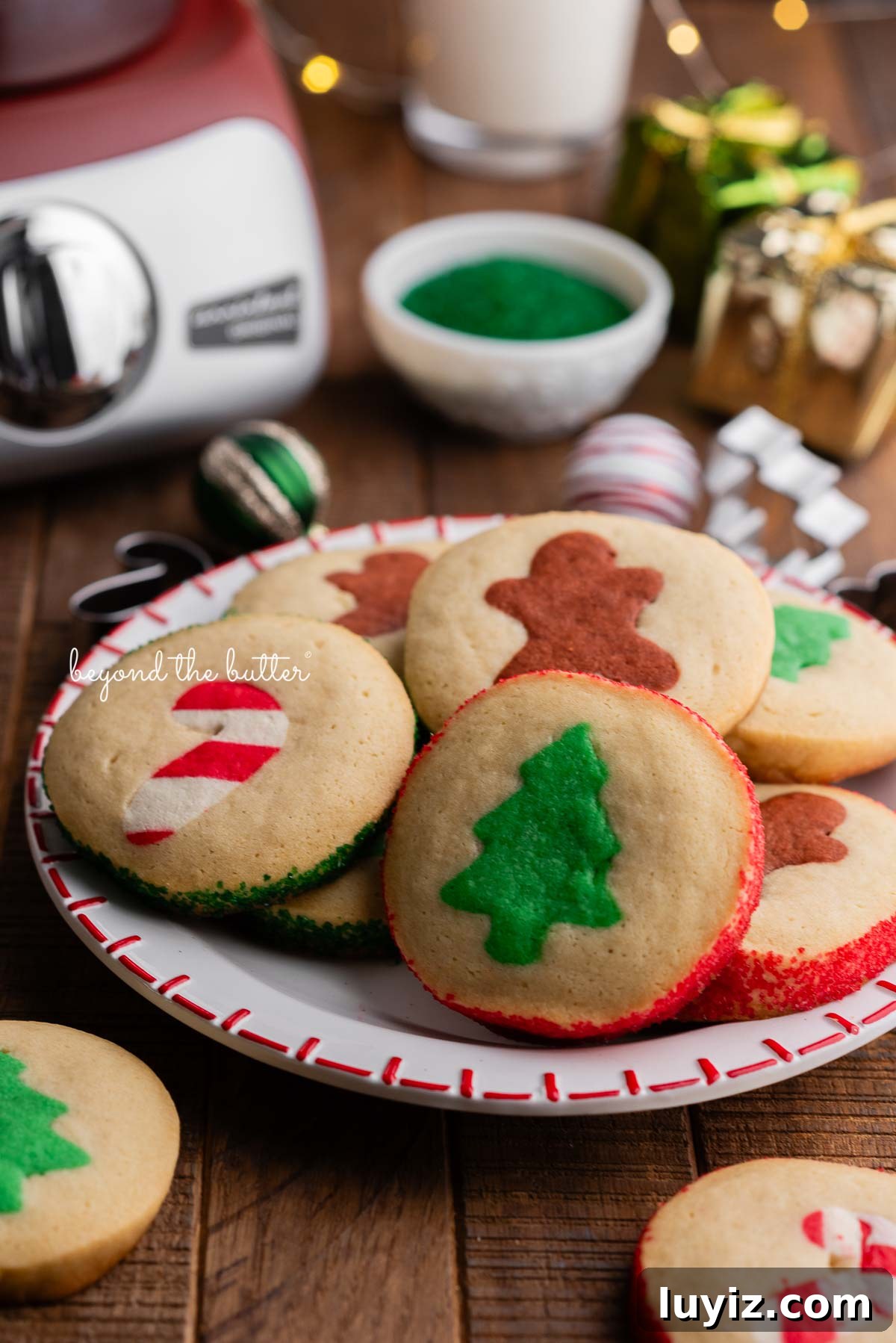 Festive Slice-and-Bake Cookies for Holiday Baking 3 A red and white holiday plate full of slice and bake cookies surrounded by more cookies, ornaments, glass of milk, sparkling sugar, and a rustic maroon colored Ankarsrum Assistent Original mixer on a dark wooden background.