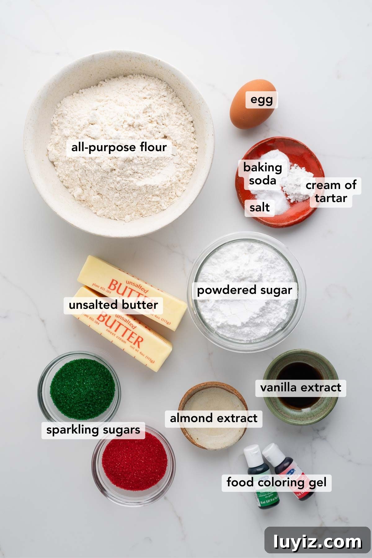 Festive Slice-and-Bake Cookies for Holiday Baking 4 Ingredients in various size bowls and plates labeled as all purpose flour, egg, baking soda, cream of tartar, salt, unsalted butter, powdered sugar, sparkling sugars, almond extract, vanilla extract, and food coloring gel on a white marbled background.