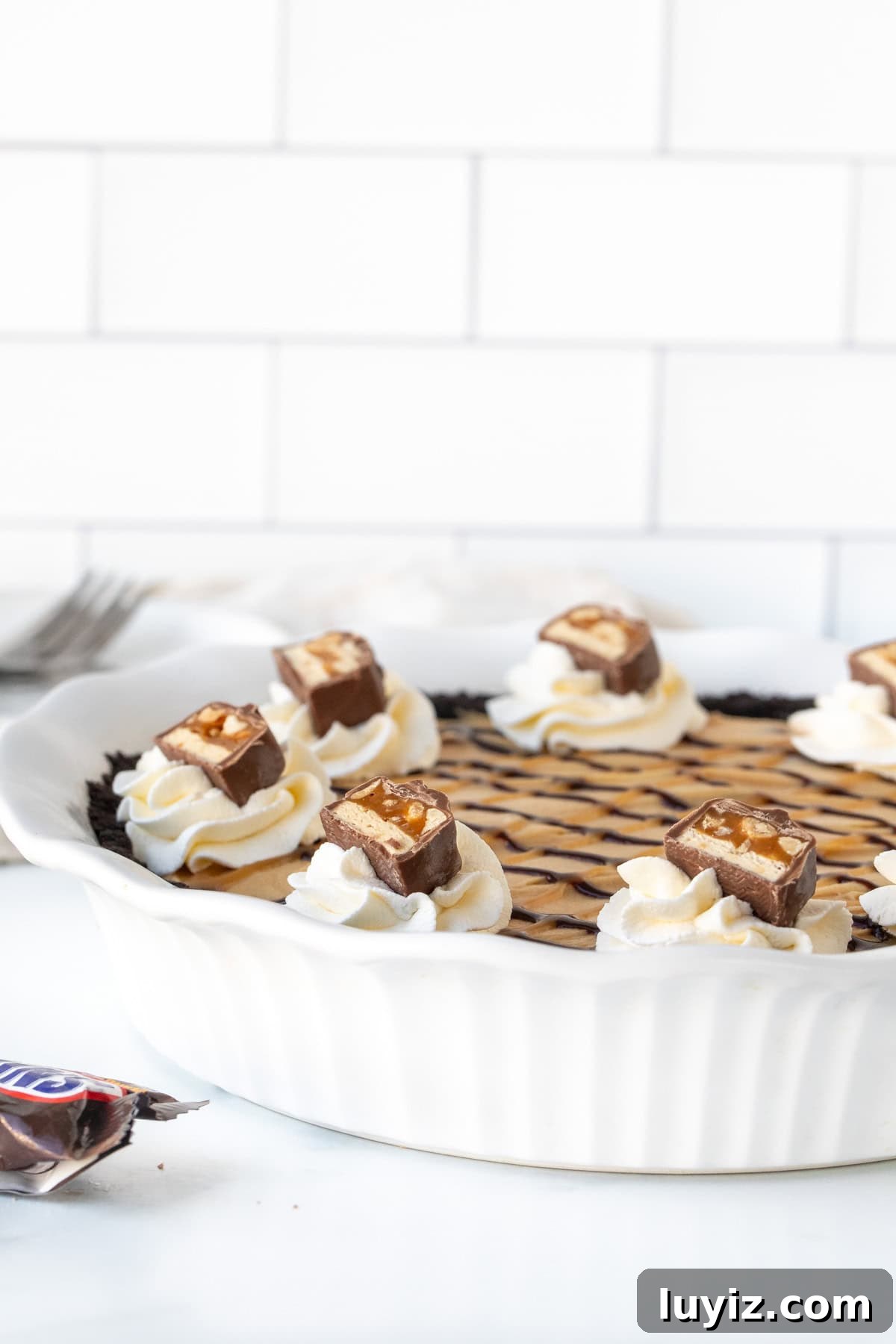 Decadent Snickers Pie Recipe — Chocolate Caramel Peanut Tart 8 Snickers bar pie in a white pie plate topped with whipped cream and chopped Snickers