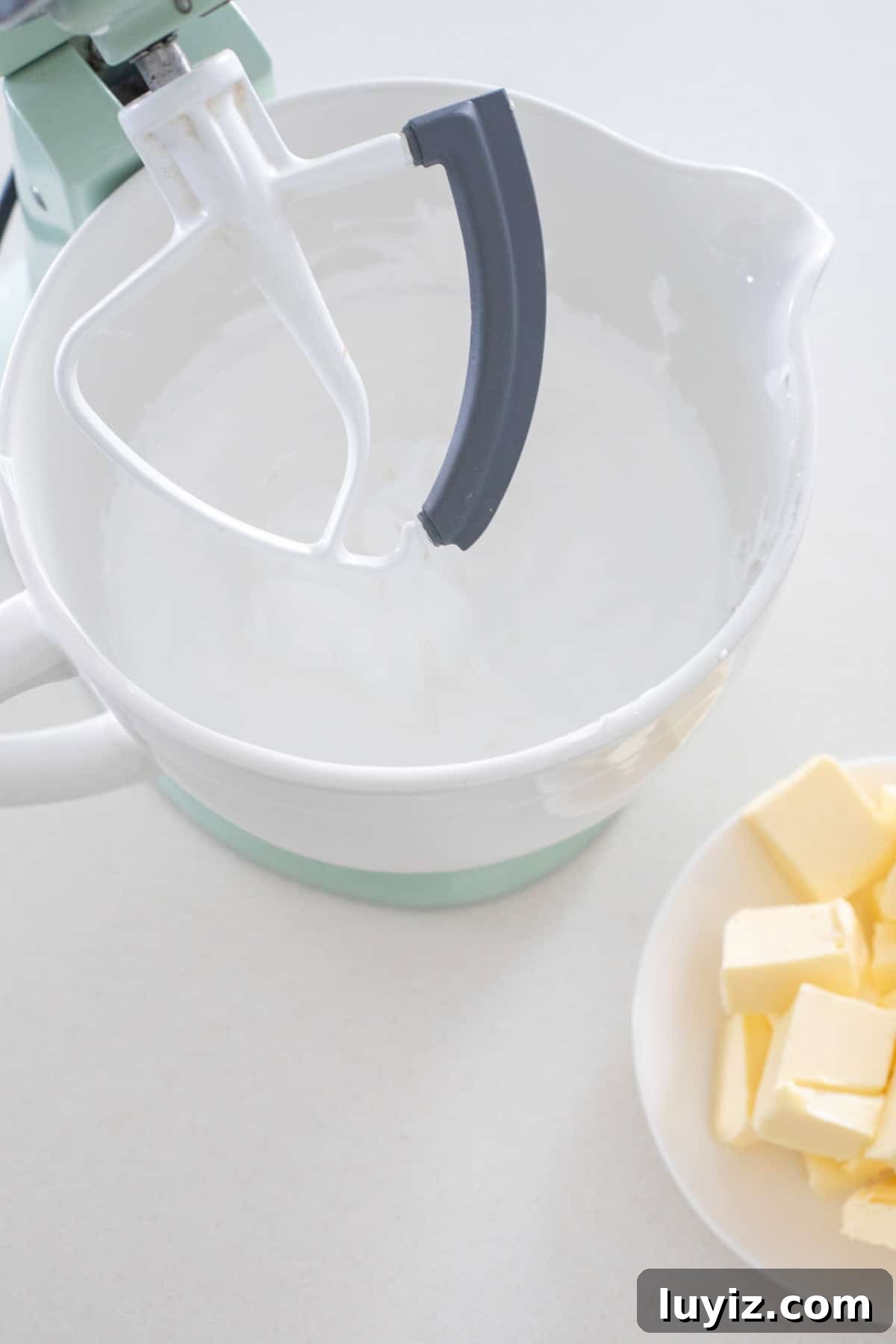 Swiss Meringue Buttercream Recipe: Smooth, Silky Frosting Guide 5 Stand mixer with paddle attachment with bowl of meringue and plate of cubed butter.