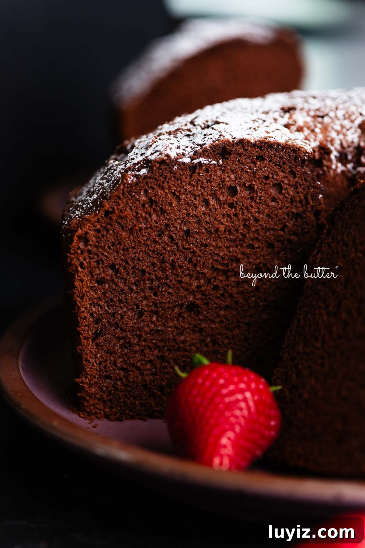 Light and Airy Chocolate Chiffon Cake Recipe 2 Chocolate chiffon cake sliced with whole strawberries placed on and in front of a dark purple ceramic plate on a black background.