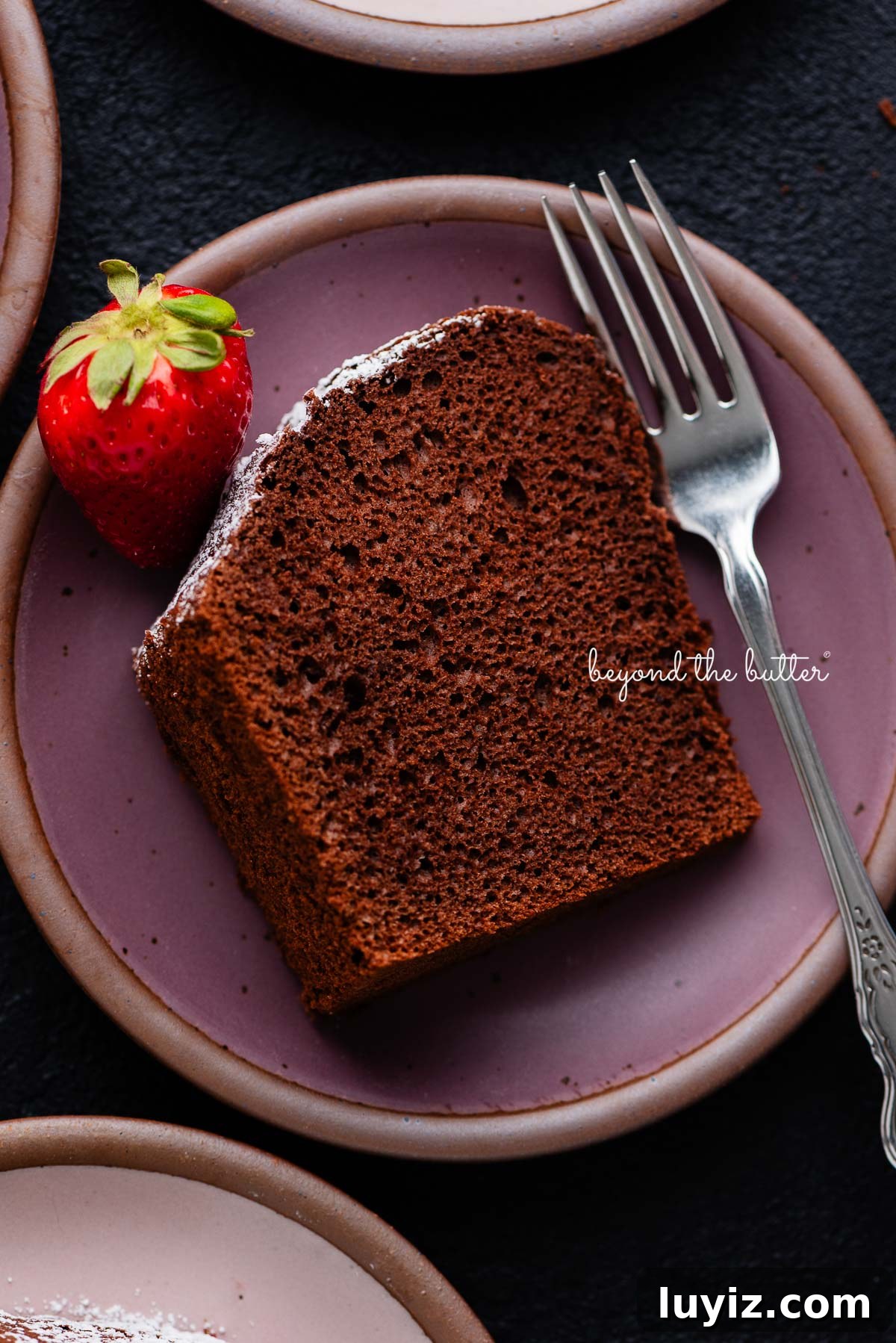 Light and Airy Chocolate Chiffon Cake Recipe 10 Slices of chocolate chiffon cake on small dark purple and light pink ceramic dessert plates with a fork and strawberry placed on each one on a black background.