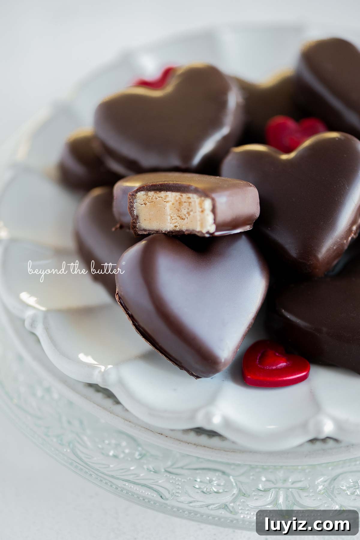 Chocolate-Covered Peanut Butter Heart Truffles 8 Chocolate covered peanut butter hearts on a white ceramic dessert plate on white background with bite taken out of center peanut butter heart.