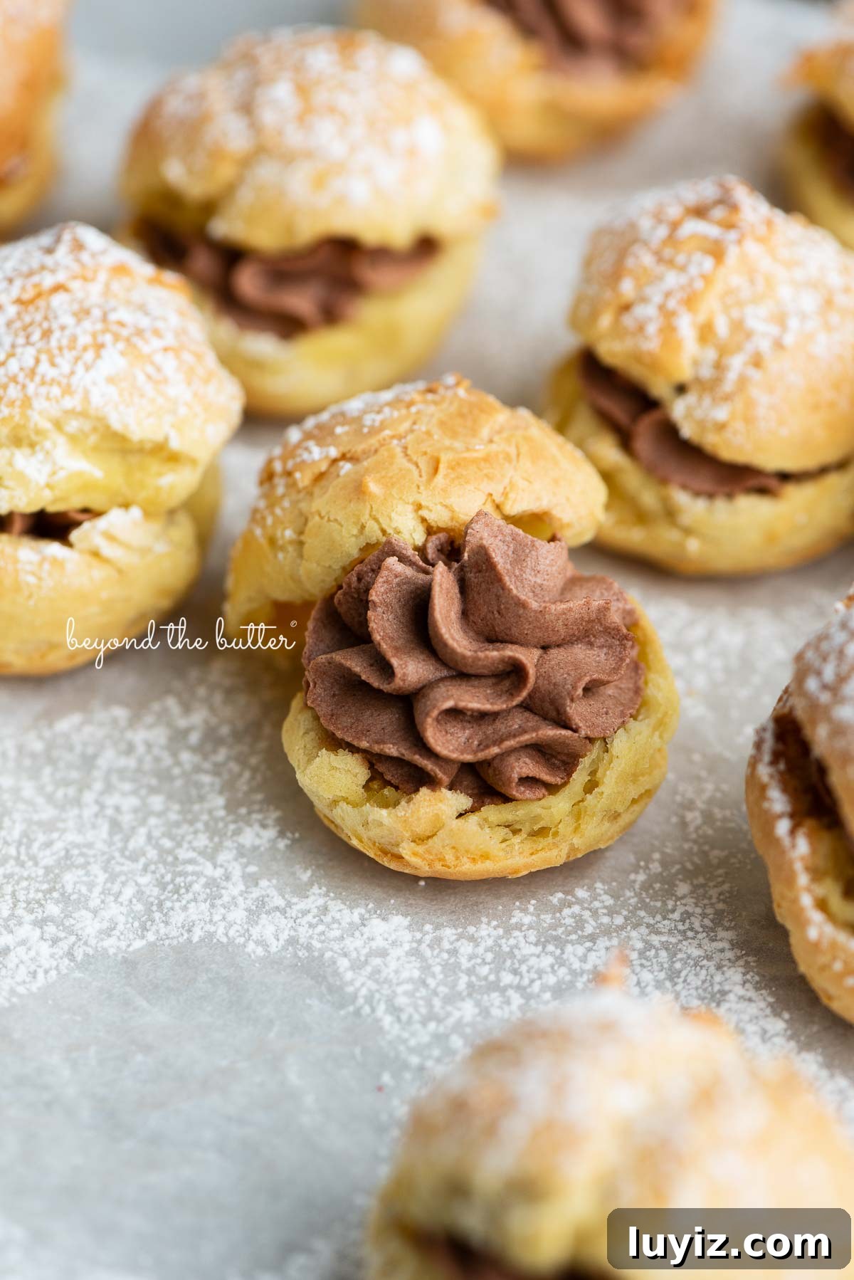 How to Make Classic Cream Puffs: Puff Pastry Filled with Vanilla Cream 2 Cream puffs filled with chocolate whipped cream and dusted with powdered sugar on a parchment paper lined baking sheet.