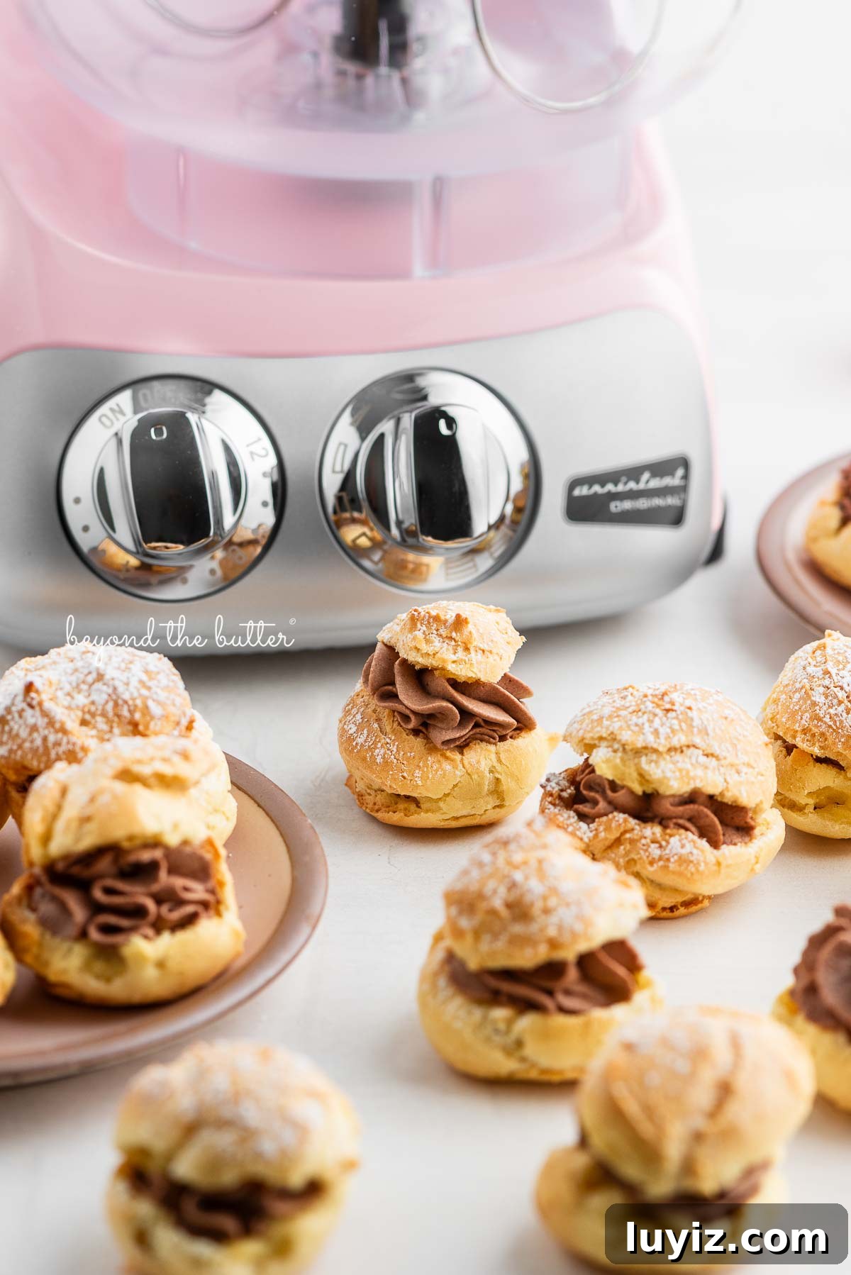 How to Make Classic Cream Puffs: Puff Pastry Filled with Vanilla Cream 3 Cloudy pink Ankarsrum mixer with cream puffs filled with chocolate whipped cream and dusted with powdered sugar placed around it on pink dessert plates and white marbled background.