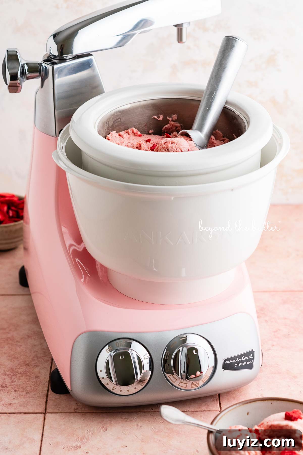 Creamy Homemade Strawberry Ice Cream Recipe You’ll Love 3 Cloudy pink Ankarsrum Assistent Original Mixer and ice cream maker attachment containing homemade strawberry ice cream with a bowl of ice cream and freeze-dried strawberries around it on a pink title background.
