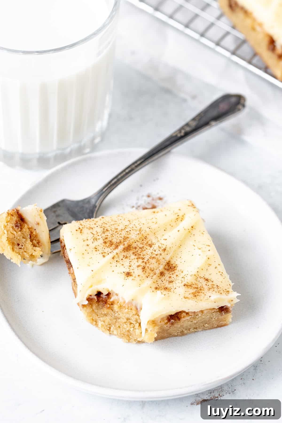 Cinnamon Roll Blondies Recipe — Gooey Swirl Dessert Bars 6 Cinnamon roll blondie with cream cheese frosting on a plate with a bite taken out by a fork.