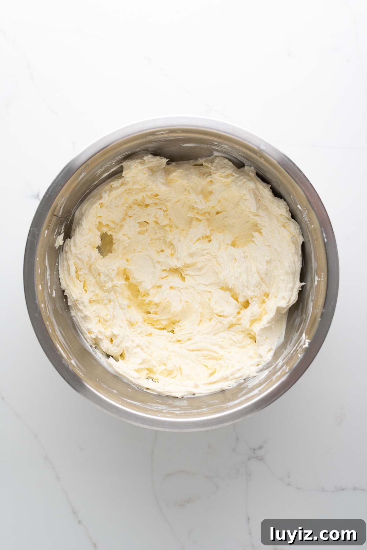 Creamy Pumpkin Cheesecake Recipe for Fall Desserts 11 Whipped cream cheese in a stainless steel mixing bowl on a white marbled background.