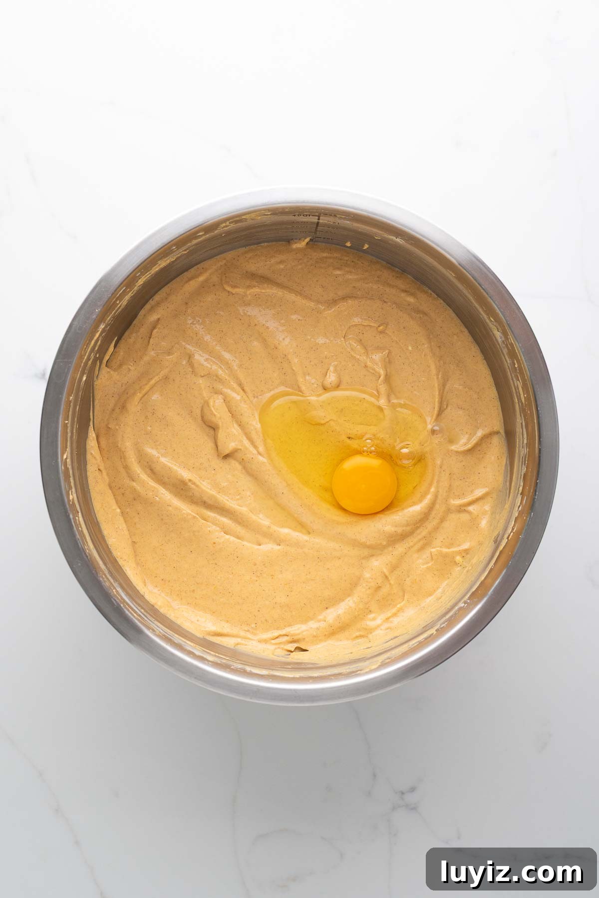 Creamy Pumpkin Cheesecake Recipe for Fall Desserts 13 Egg added to pumpkin cheesecake batter in a stainless steel mixing bowl on a white marbled background.