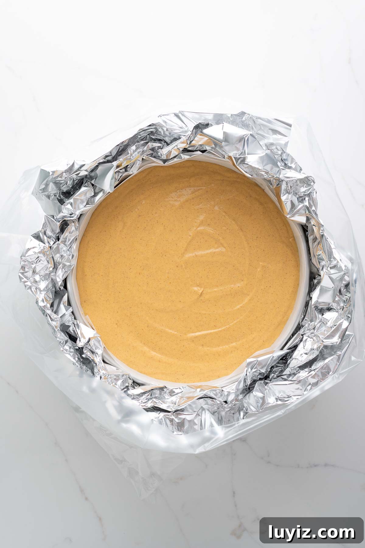 Creamy Pumpkin Cheesecake Recipe for Fall Desserts 14 Pumpkin cheesecake in a parchment paper lined springform pan wrapped in aluminum foil and a slow cooker bag on a white marbled background.