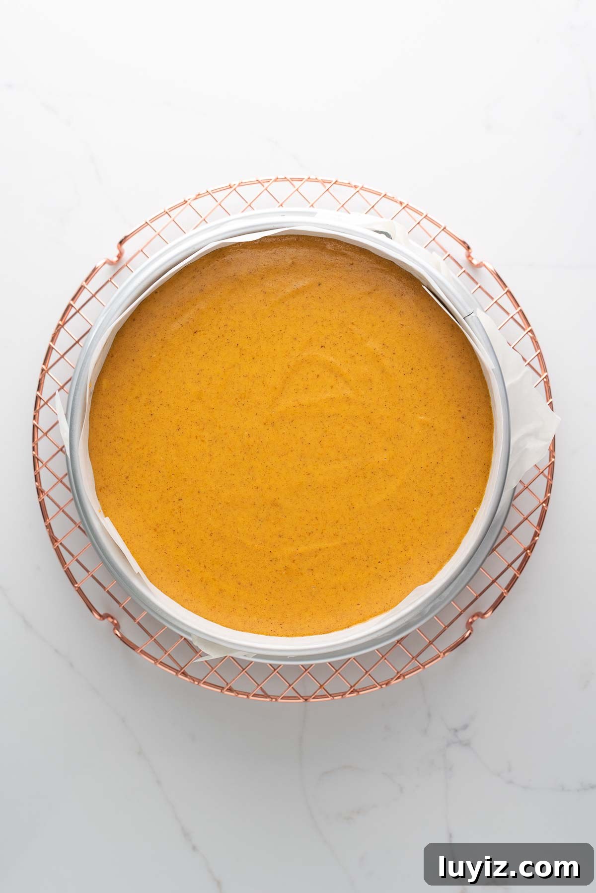 Creamy Pumpkin Cheesecake Recipe for Fall Desserts 17 Cooling a pumpkin cheesecake on a round rose gold colored cooling rack on a white marbled background.