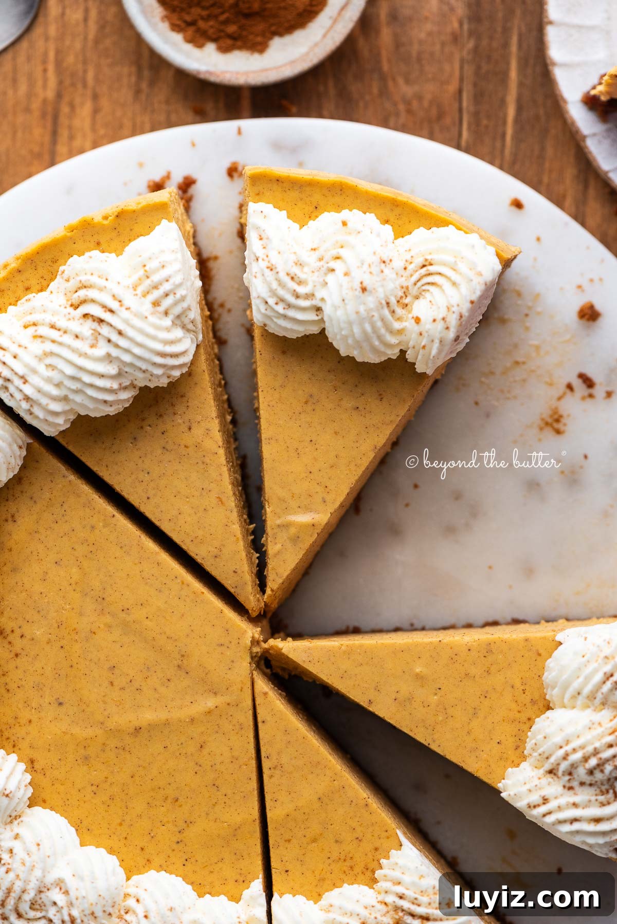 Creamy Pumpkin Cheesecake Recipe for Fall Desserts 19 Slices of pumpkin cheesecake topped with piped on whipped cream on a marbled cake plate and dark wood background.