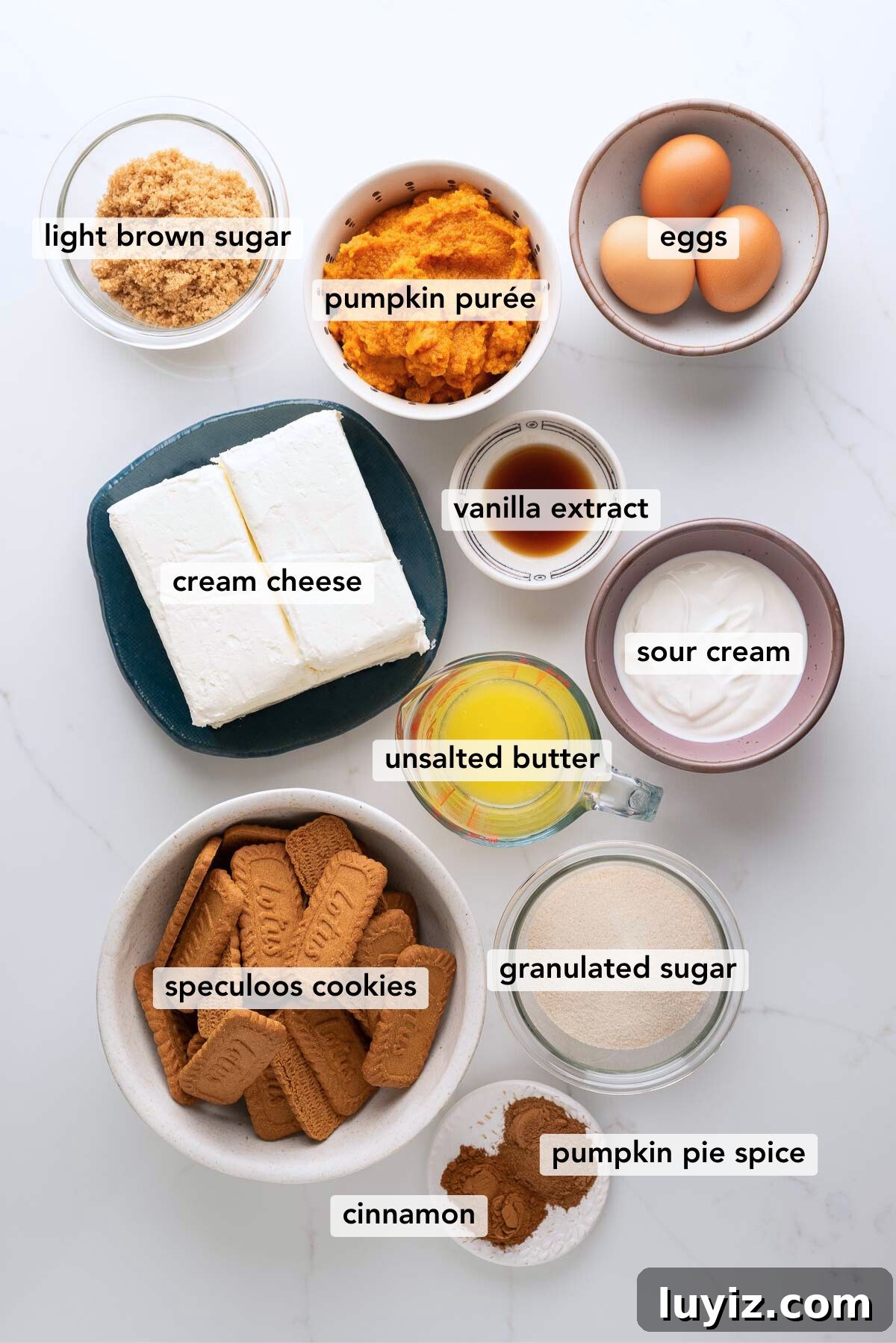 Creamy Pumpkin Cheesecake Recipe for Fall Desserts 4 Ingredients in various size bowls labeled as light brown sugar, pumpkin puree, eggs, cream cheese, vanilla extract, unsalted butter, sour cream, speculoos cookies,granulated sugar, cinnamon, and pumpkin pie spice on a white marbled background.