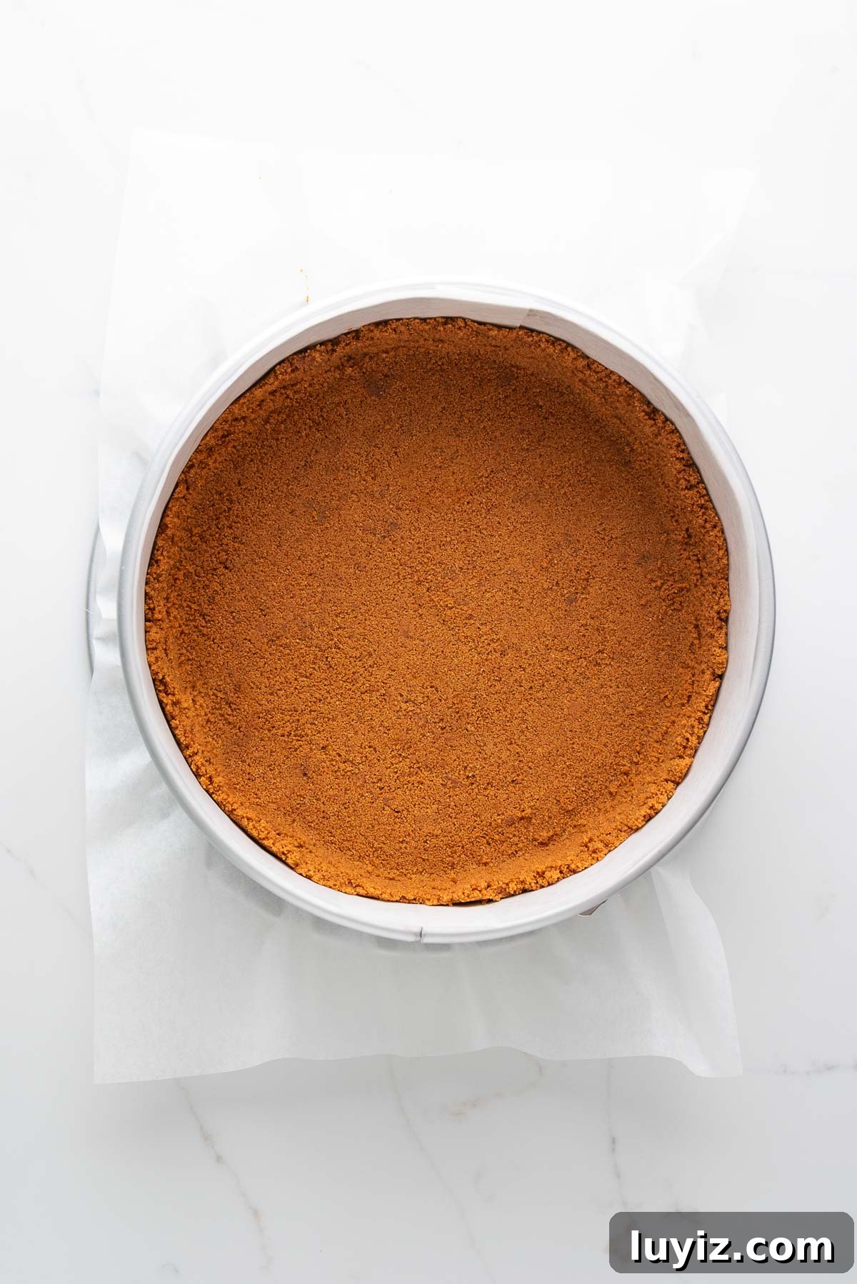 Creamy Pumpkin Cheesecake Recipe for Fall Desserts 9 Baked speculoos cookie crust in a parchment paper lined springform pan on a white marbled background.