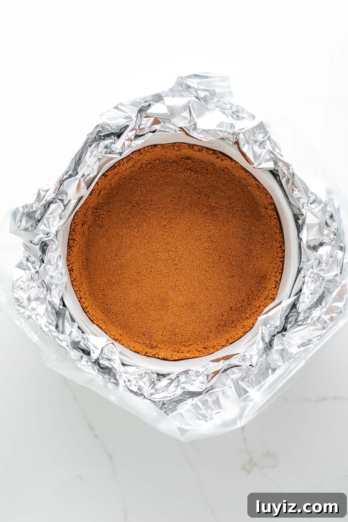 Creamy Pumpkin Cheesecake Recipe for Fall Desserts 10 Biscoff cookie crust in a parchment paper lined springform pan wrapped with sheets of aluminum foil and a slow cooker liner.