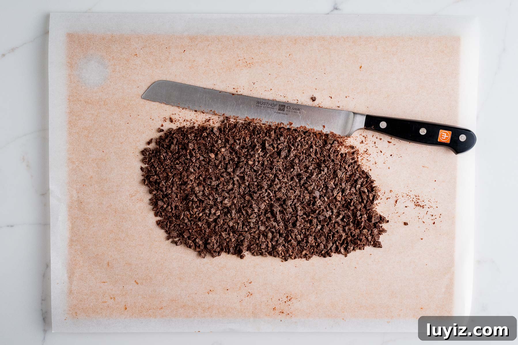 Chocolate Croissant Loaf Recipe: Flaky Chocolate Swirl Bread 12 Finely chopped dark chocolate on a parchment paper lined cutting board with a serrated knife on a white marbled background.
