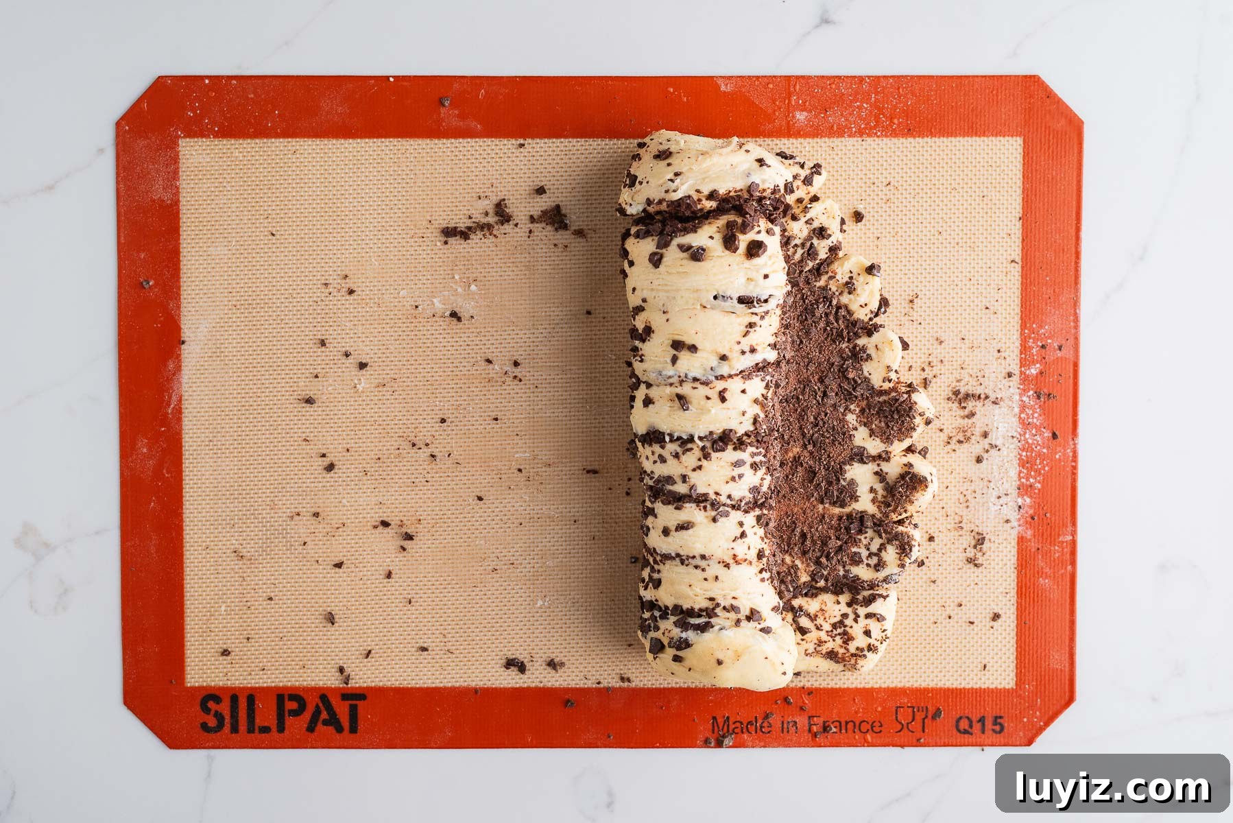 Chocolate Croissant Loaf Recipe: Flaky Chocolate Swirl Bread 14 Rolling up the chocolate croissant dough loaf on a silicone baking sheet on a white marbled background.
