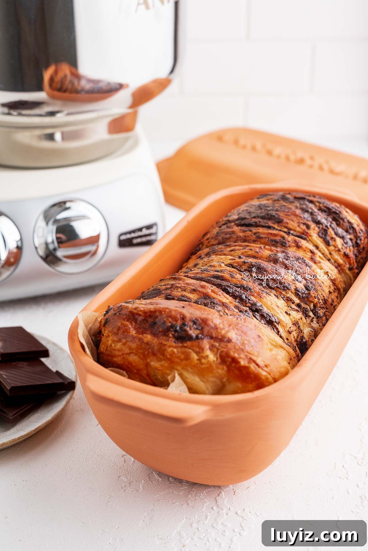 Chocolate Croissant Loaf Recipe: Flaky Chocolate Swirl Bread 18 A chocolate croissant loaf placed in a parchment paper lined Ankasrum Clay Baker and next to a plate of dark chocolate and a cream colored Ankarsrum Assistent Original Mixer on a white background.