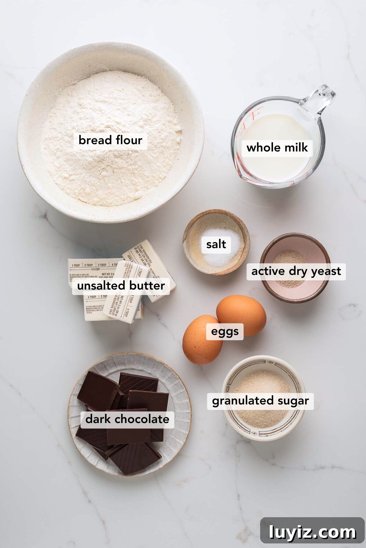 Chocolate Croissant Loaf Recipe: Flaky Chocolate Swirl Bread 5 Ingredients in various size bowls labeled as bread flour, whole milk, unsalted butter, salt, active dry yeast, eggs, dark chocolate, and granulated sugar on a white marbled background.