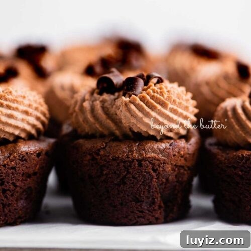Fudgy brownie bites topped with chocolate mascarpone whipped cream and chocolate curls placed together on a parchment paper lined baking sheet.