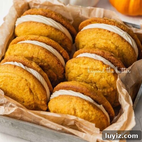 Six pumpkin whoopie pies placed in a brown parchment paper lined metal baking tin with mini orange and white pumpkins placed around it on a white background.