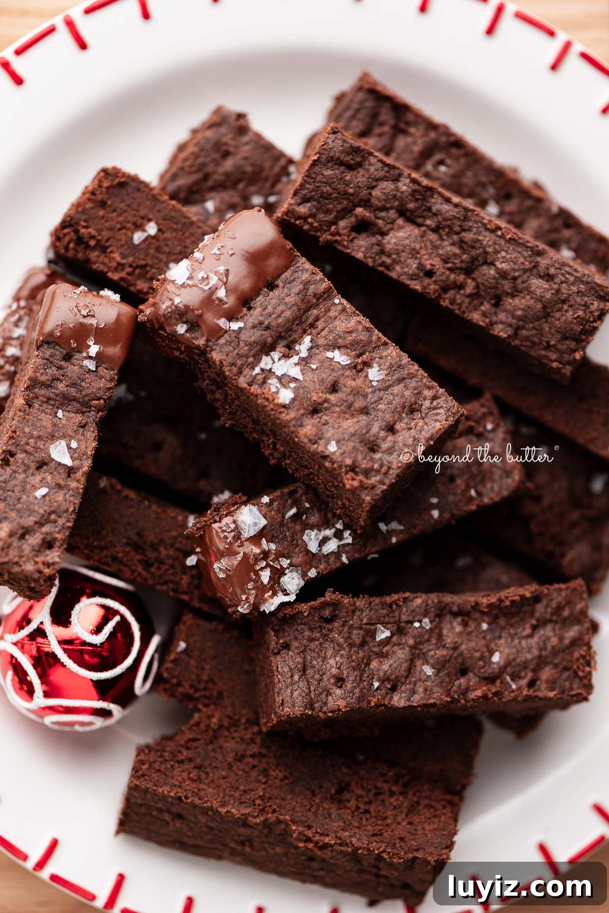 Classic Chocolate Shortbread Cookies Recipe for Homemade Treats 14 Sliced chocolate shortbread biscuits sprinkled with flaky sea salt flakes and placed on a round red and white holiday-themed dessert plate surrounded by miniature ball ornaments on a wood surface.