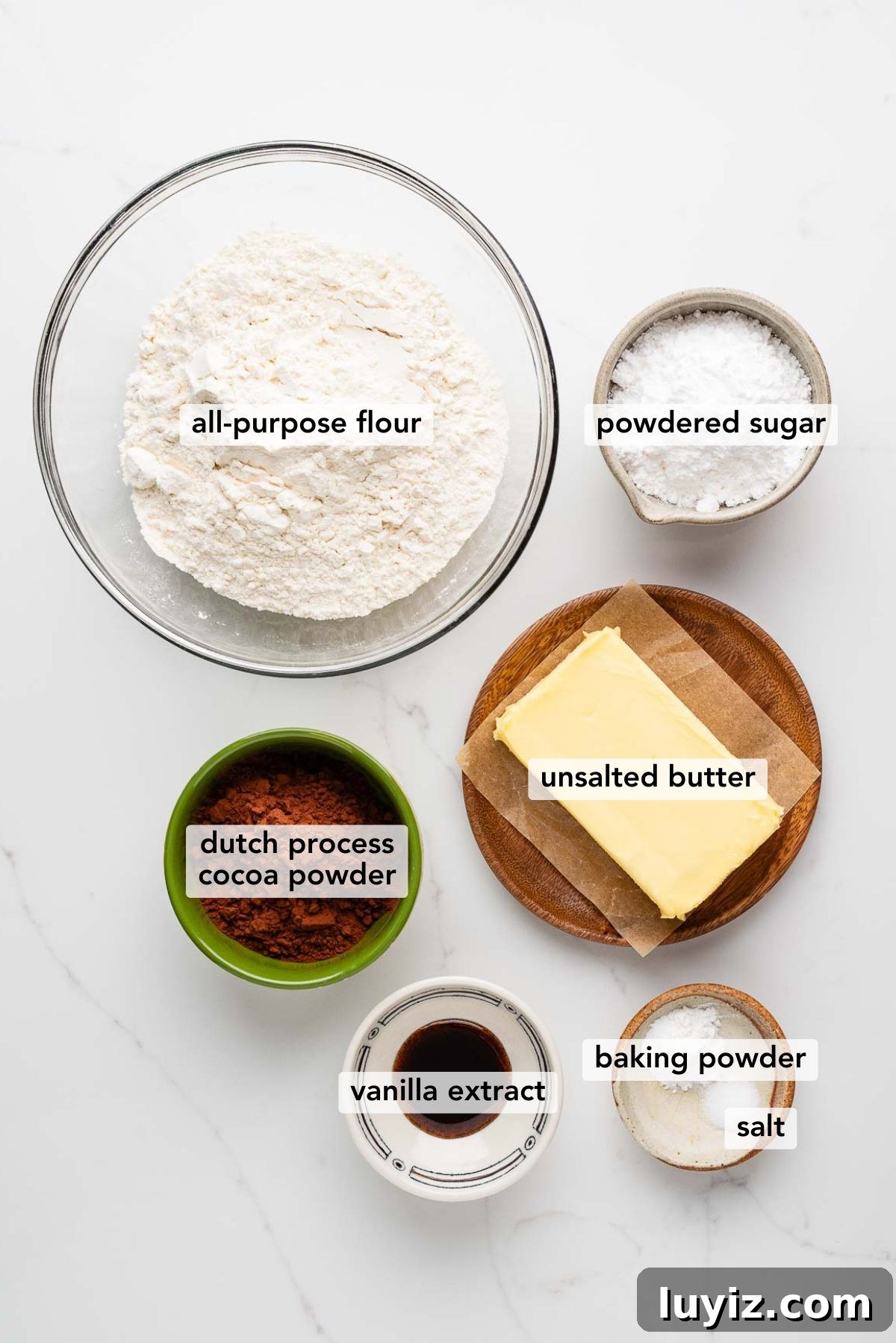 Classic Chocolate Shortbread Cookies Recipe for Homemade Treats 4 Ingredients in various size bowls labeled as all purpose flour, powdered sugar, dutch process cocoa powder, unsalted butter, vanilla extract, baking powder, and salt on a white marbled background.