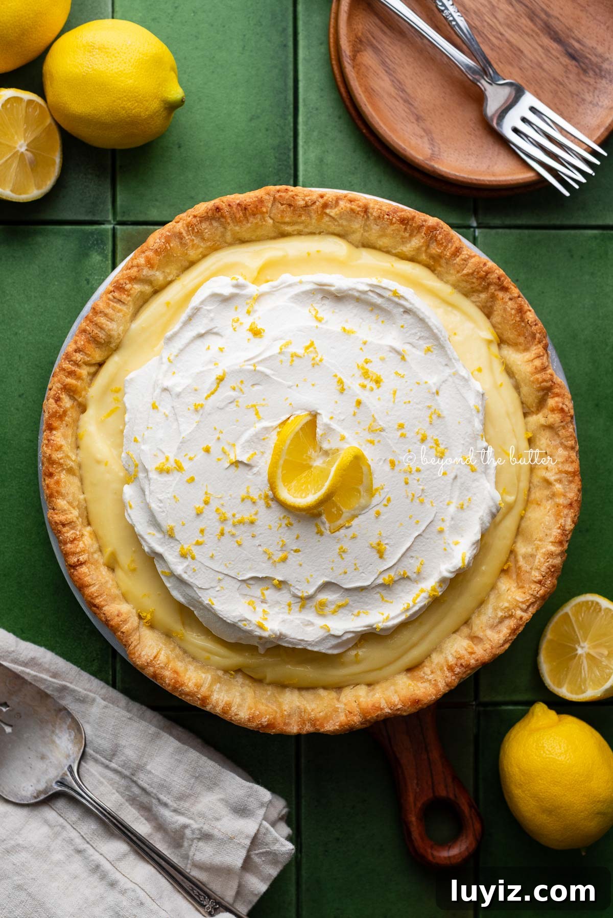 Lemon Cream Pie Recipe: Silky No-Bake Dessert Tutorial 2 Lemon cream pie topped with fresh whipped cream and lemon zest centered on a green tiled background surrounded by lemons, wood dessert plates, forks, napkin, and pie server.