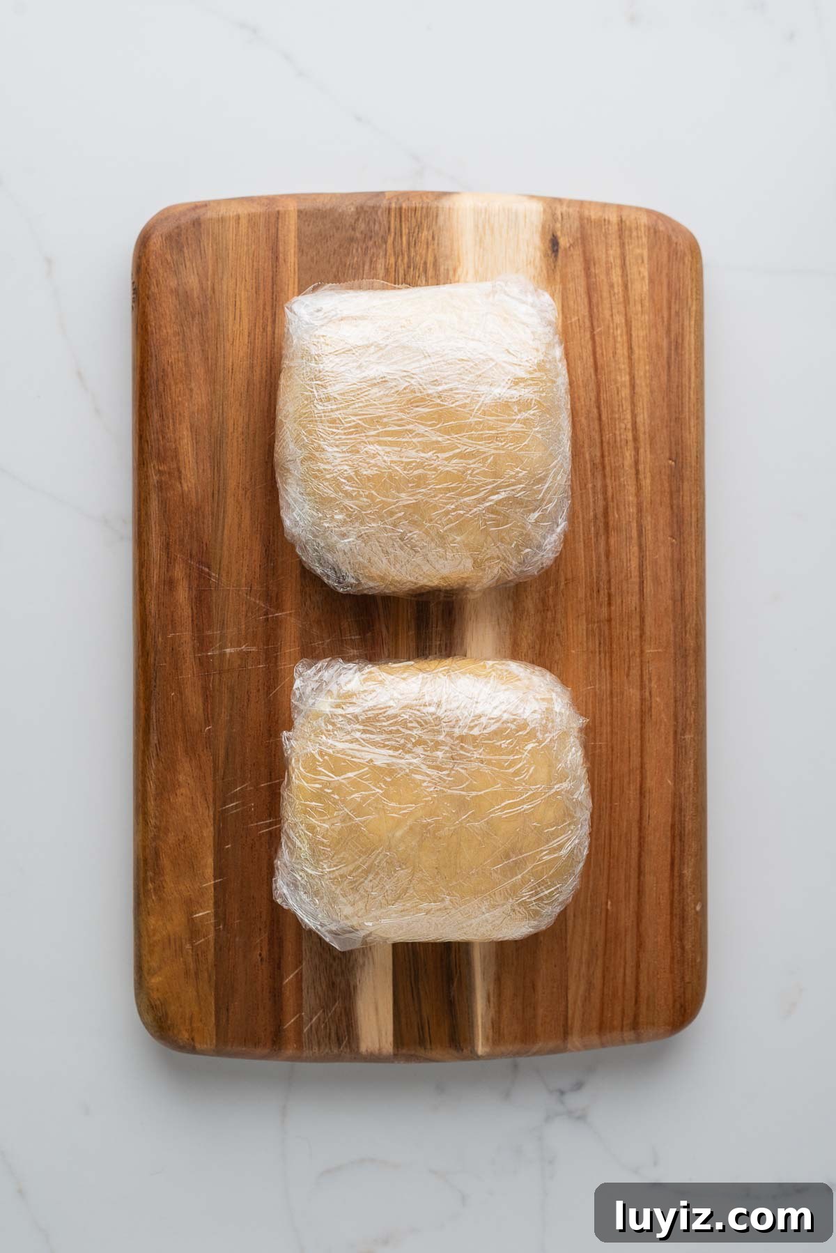 Lemon Cream Pie Recipe: Silky No-Bake Dessert Tutorial 11 Two pie dough discs double wrapped in plastic wrap placed on a wood cutting board on a white marbled background.