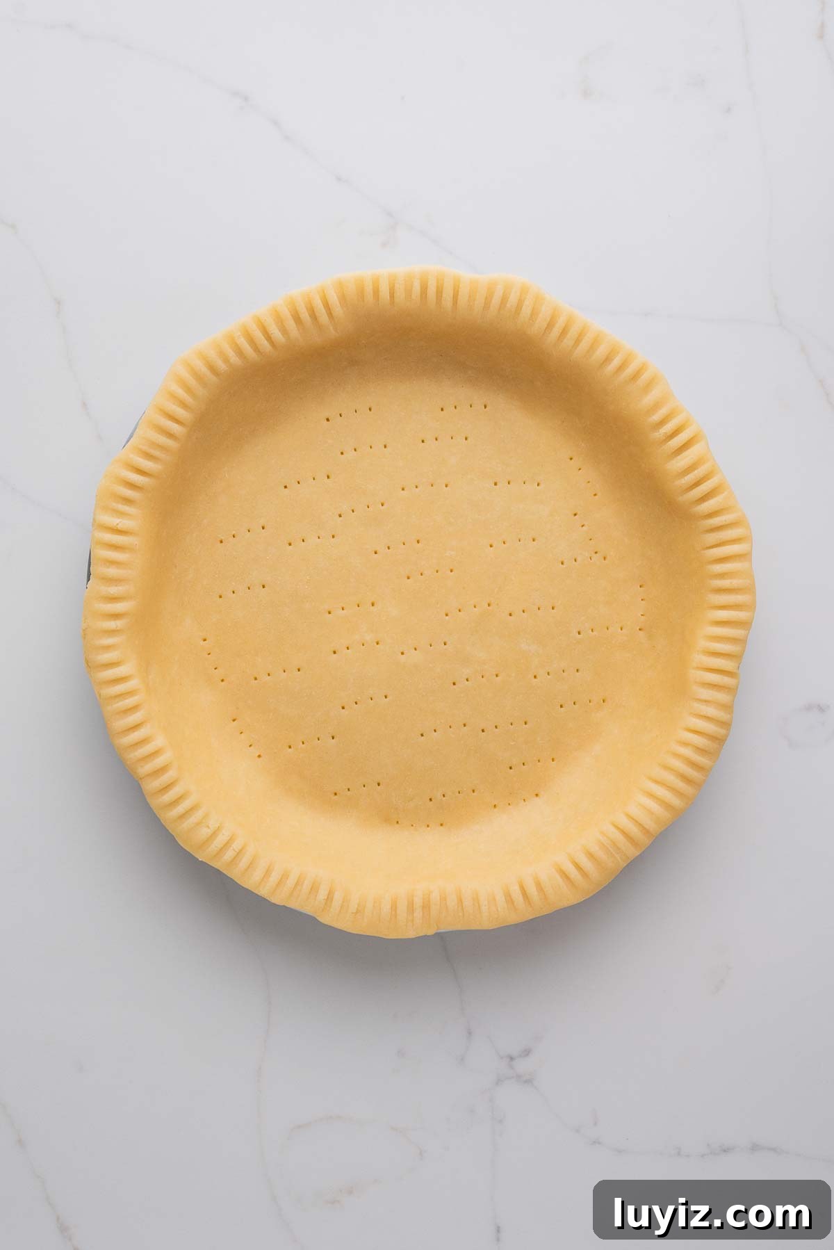 Lemon Cream Pie Recipe: Silky No-Bake Dessert Tutorial 13 Rolled out pie dough placed in a metal pie tin that's been trimmed, edges crimped and bottom docked with a fork on a white marbled background.