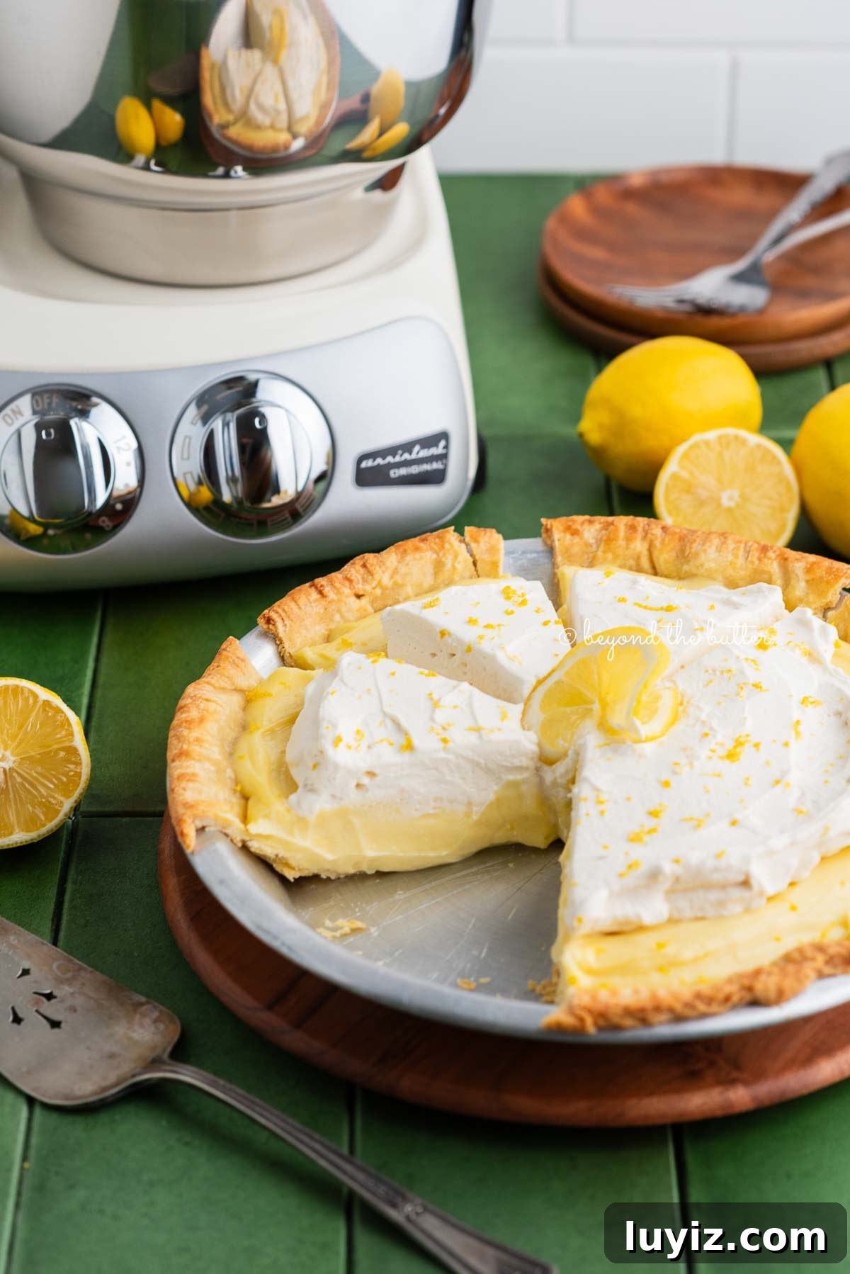 Lemon Cream Pie Recipe: Silky No-Bake Dessert Tutorial 4 Sliced lemon sour cream pie on a wooden pie serving board next to a creme light colored ankarsrum assistent original mixer, lemons, pie server, and dessert plates.