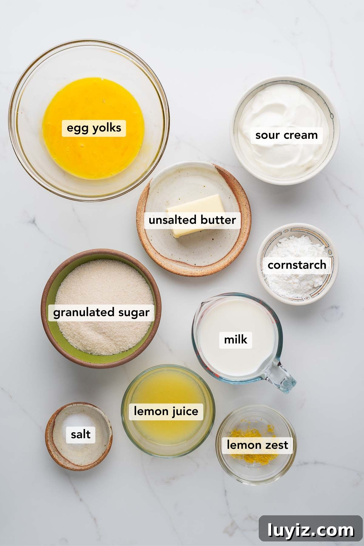 Lemon Cream Pie Recipe: Silky No-Bake Dessert Tutorial 7 Ingredients in various size bowls labeled as egg yolks, sour cream, unsalted butter, granulated sugar, milk, cornstarch, lemon juice, lemon zest, and salt on a white marbled background.