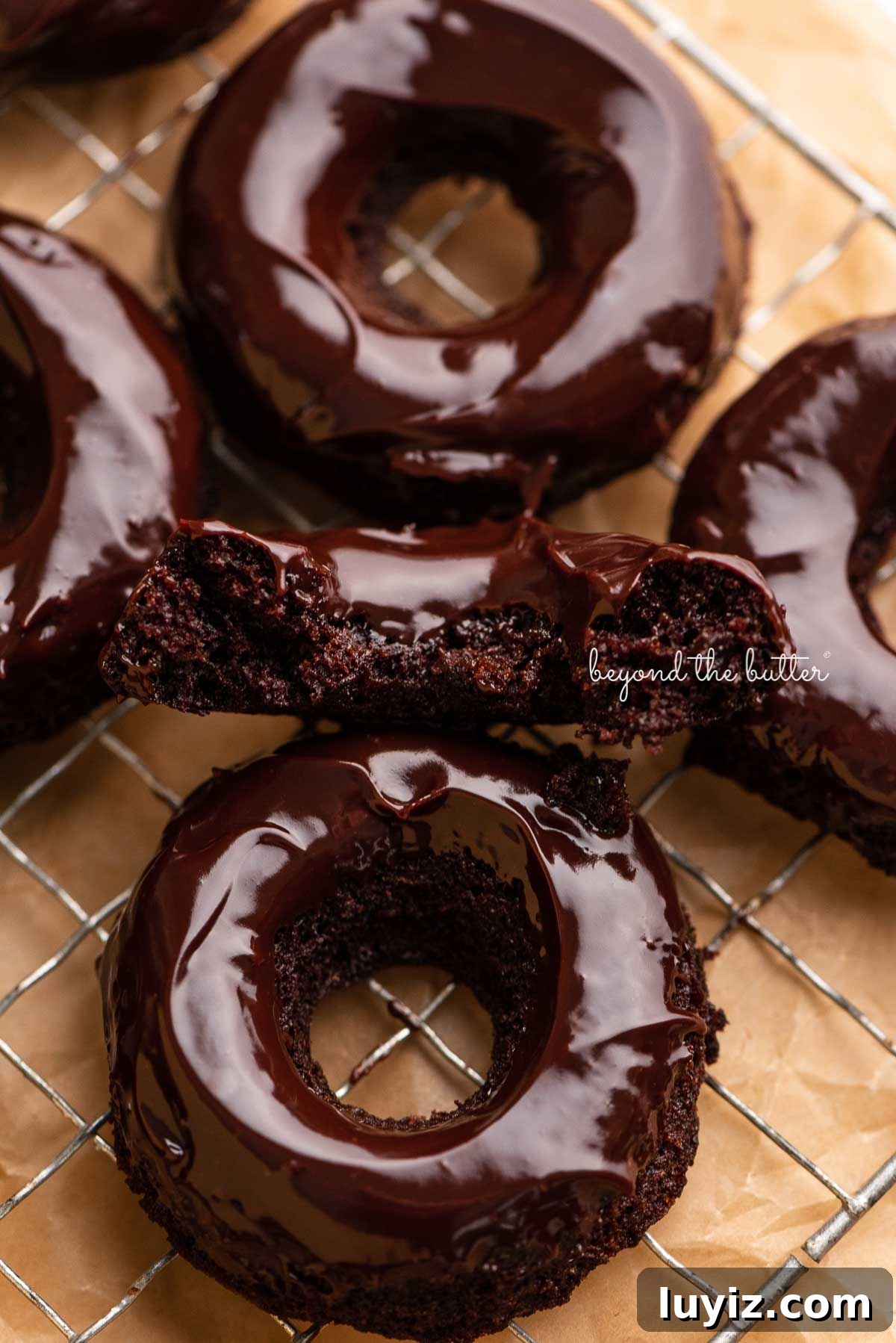 Homemade Baked Chocolate Donuts Recipe for Perfect Texture 3 Half of a homemade baked chocolate donut topped with chocolate ganache on a wire cooling rack with more donuts | © Beyond the Butter®