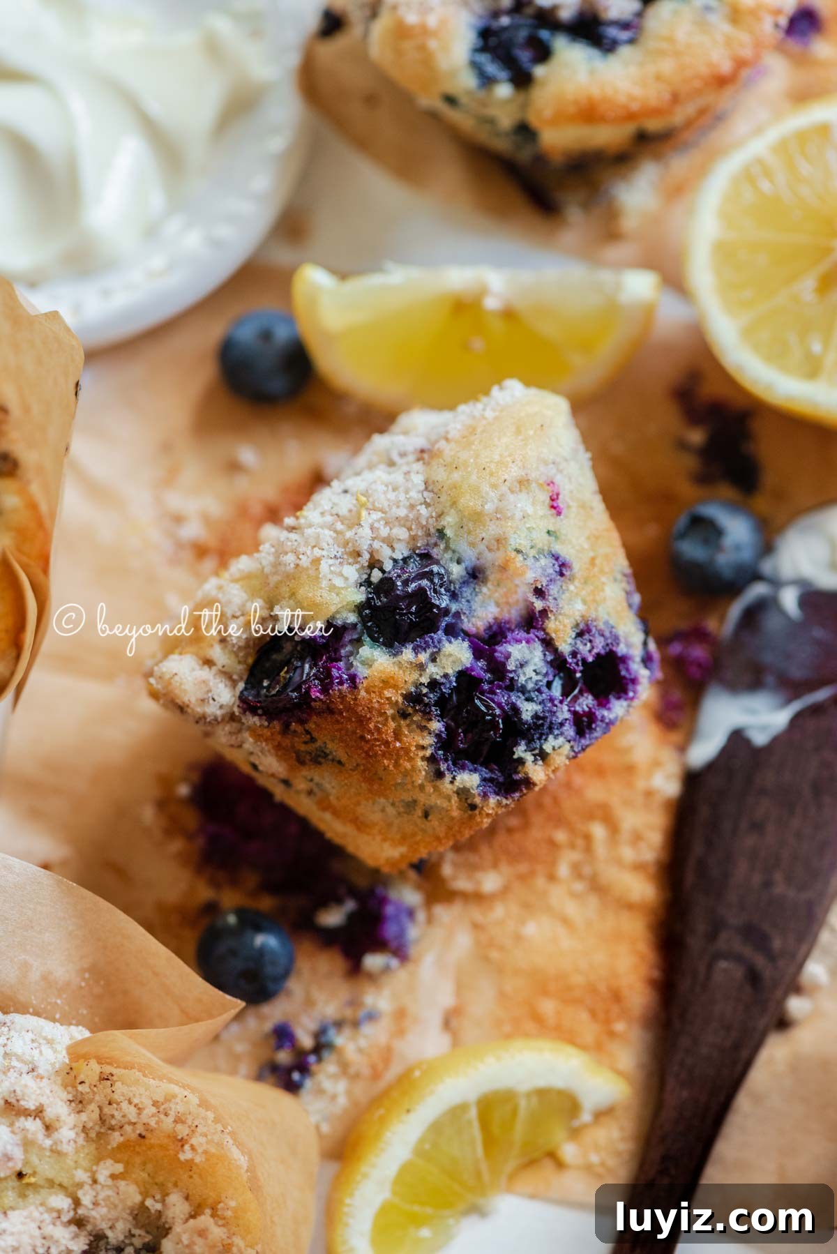 Bakery-Style Lemon Blueberry Streusel Muffins Recipe 4 Unwrapped lemon blueberry streusel muffin with small bowl of butter, lemon wedges and blueberries around it | All images © Beyond the Butter®